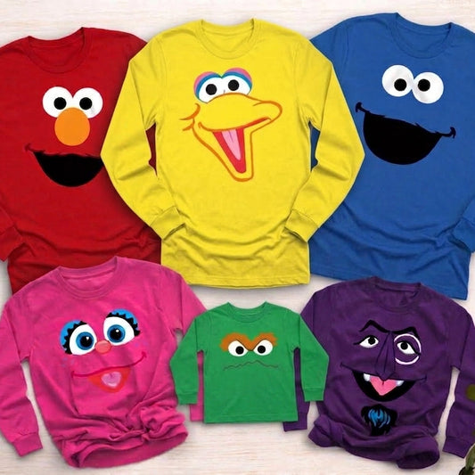 Six long-sleeve children's shirts in different colors, each printed with a Sesame Street character face, arranged on a light wood background with green leaves in the corners.