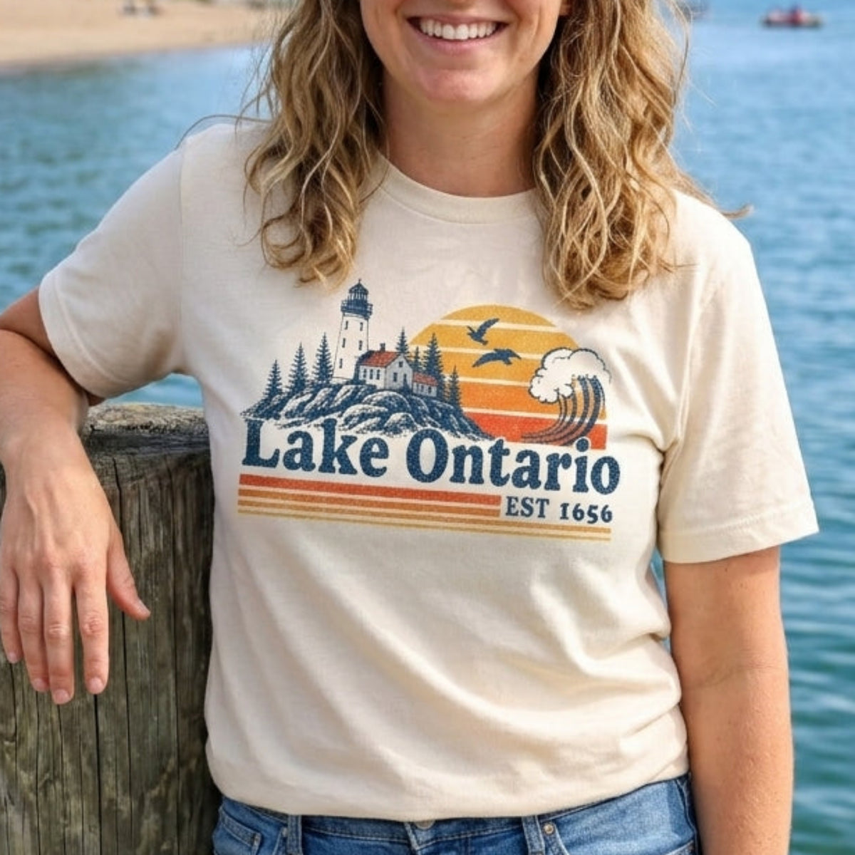 A woman smiling while leaning on a wooden dock by the water, wearing a cream-colored graphic t-shirt that reads 'Lake Ontario Est. 1650' with a sunset and lighthouse design, paired with blue jeans.