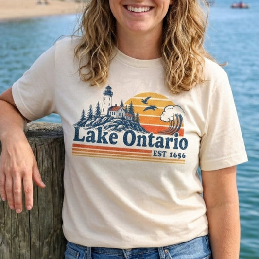 A woman smiling while leaning on a wooden dock by the water, wearing a cream-colored graphic t-shirt that reads 'Lake Ontario Est. 1650' with a sunset and lighthouse design, paired with blue jeans.