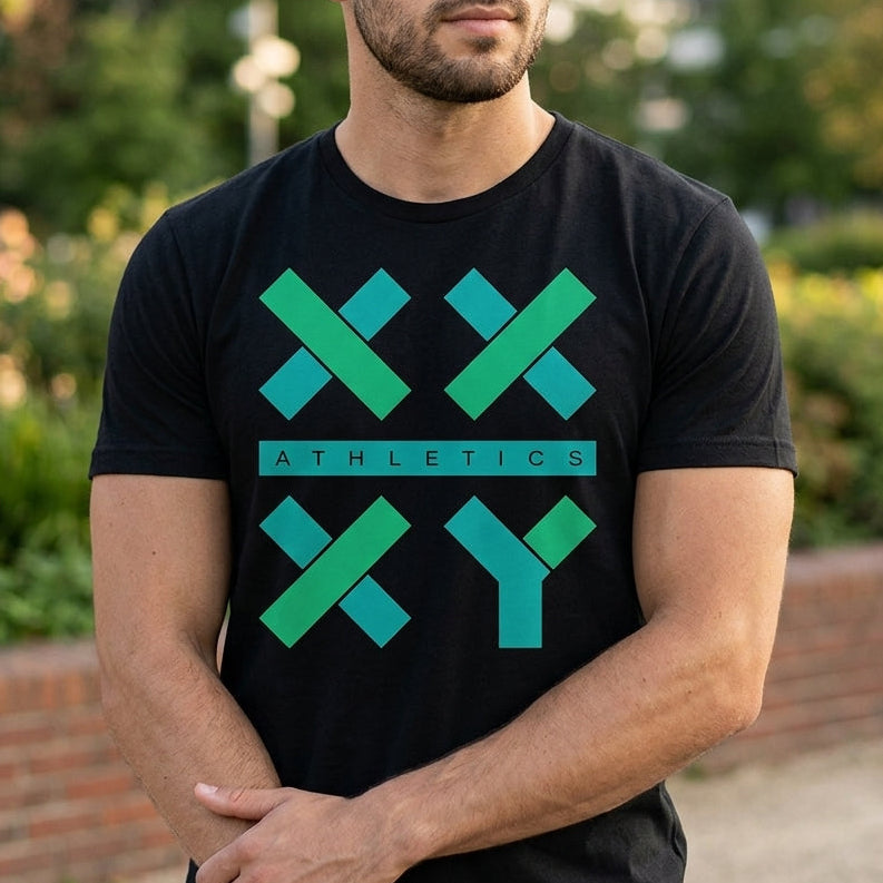 A man wearing a black short-sleeve t-shirt with a green and teal geometric 'XXY ATHLETICS' logo on the front, standing outdoors in a park-like setting with blurred greenery and a brick wall in the background.