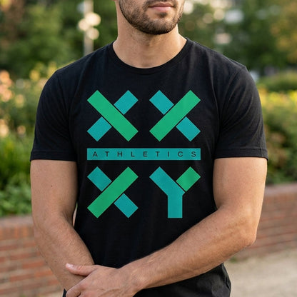 A man wearing a black short-sleeve t-shirt with a green and teal geometric 'XXY ATHLETICS' logo on the front, standing outdoors in a park-like setting with blurred greenery and a brick wall in the background.