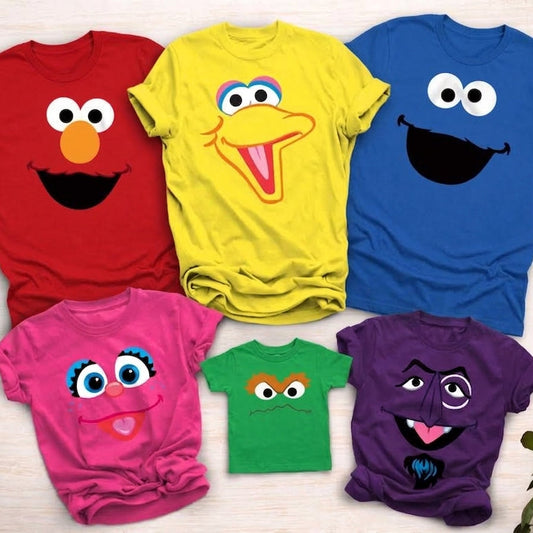 Six colorful children's t-shirts featuring Sesame Street character faces, arranged on a light wood background with green leaves in the corners. The shirts are red, yellow, blue, pink, green, and purple.