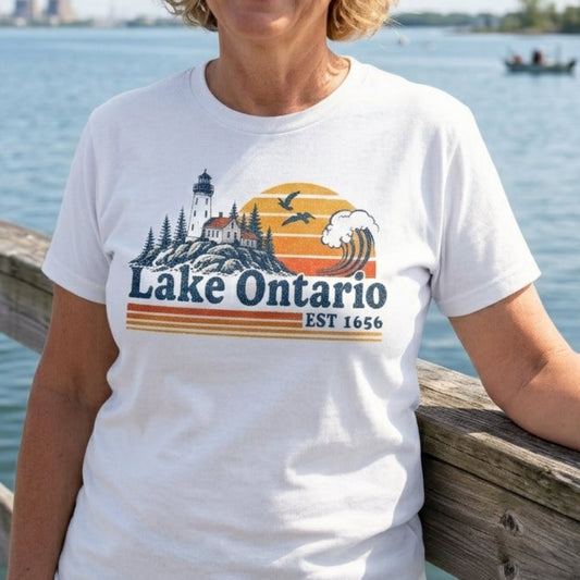 A smiling blonde woman stands on a wooden dock by the water wearing a white graphic T-shirt that reads 'Lake Ontario Est 1650' with a lighthouse, trees, and sunset design.